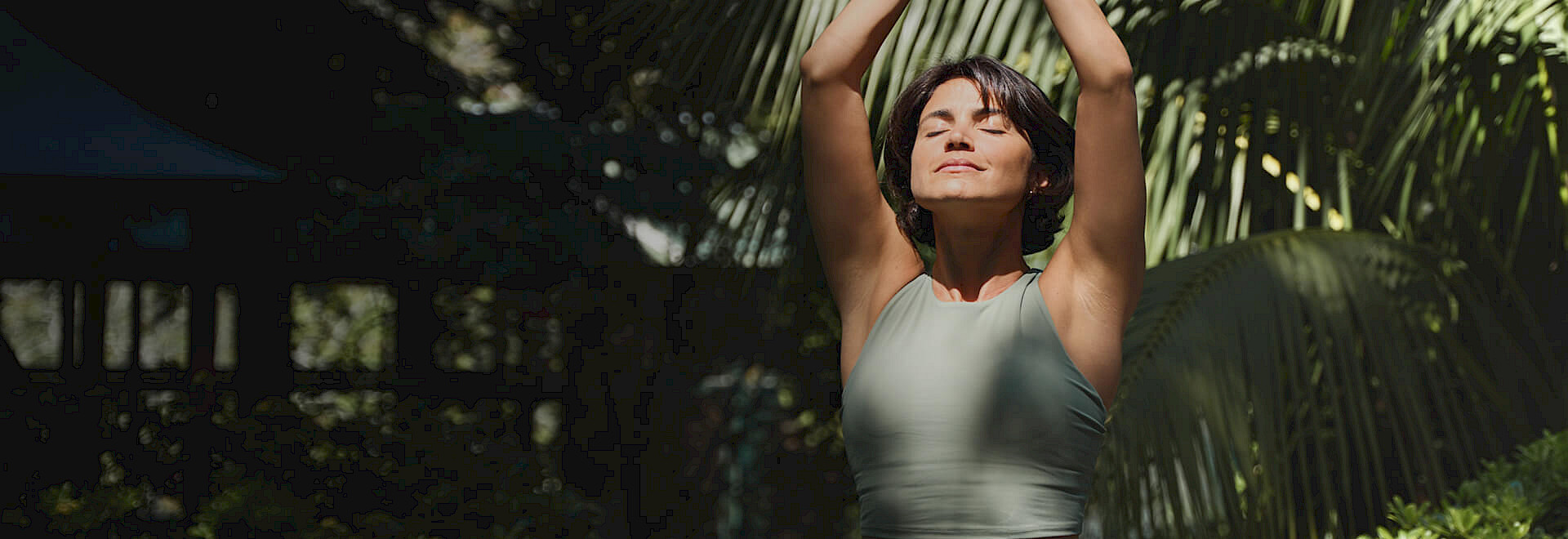 A woman in a green top practicing mindfulness with her arms raised in a lush, sunlit garden.